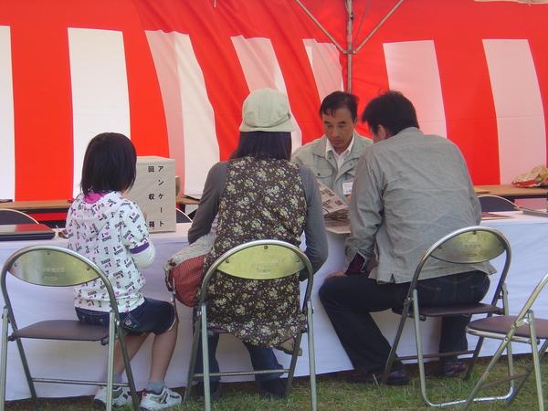 紅白幕が張られたテントの中で椅子に座り説明を聞いている親子3人の写真